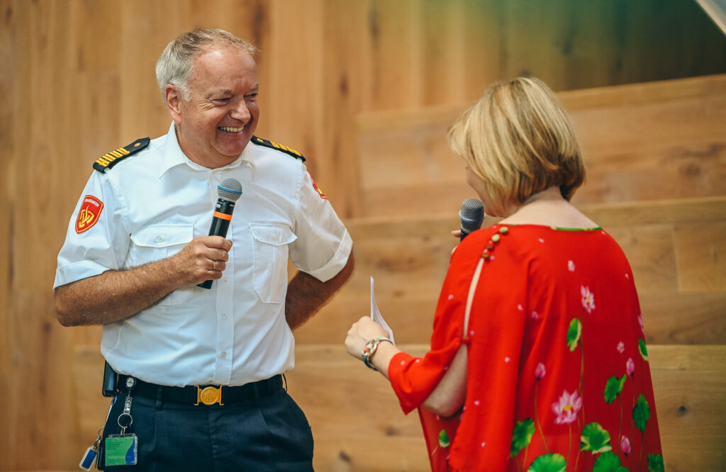 Een dagvoorzitter in een wit uniform gaat in gesprek met een vrouw in een rode jurk. Beiden houden ze een microfoon vast op een houten podium.